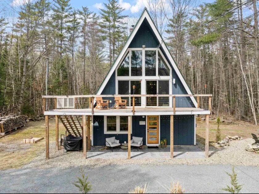 A-frame house with large front windows, elevated deck with seating, outdoor stairs, and surrounded by trees in a wooded area.