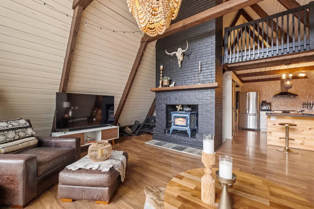 A-frame cabin living area with wood floors, a black brick fireplace, mounted bull skull, leather chair, TV, and open kitchen with loft above.
