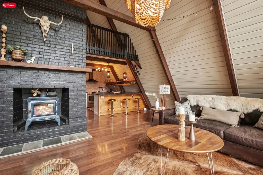 House For Sale | A Cozy Updated New Hampshire Chalet in the Mountain Lakes District. A-frame cabin interior with wood floors, black brick fireplace, wood stove, rustic decor, open kitchen, and loft area above.