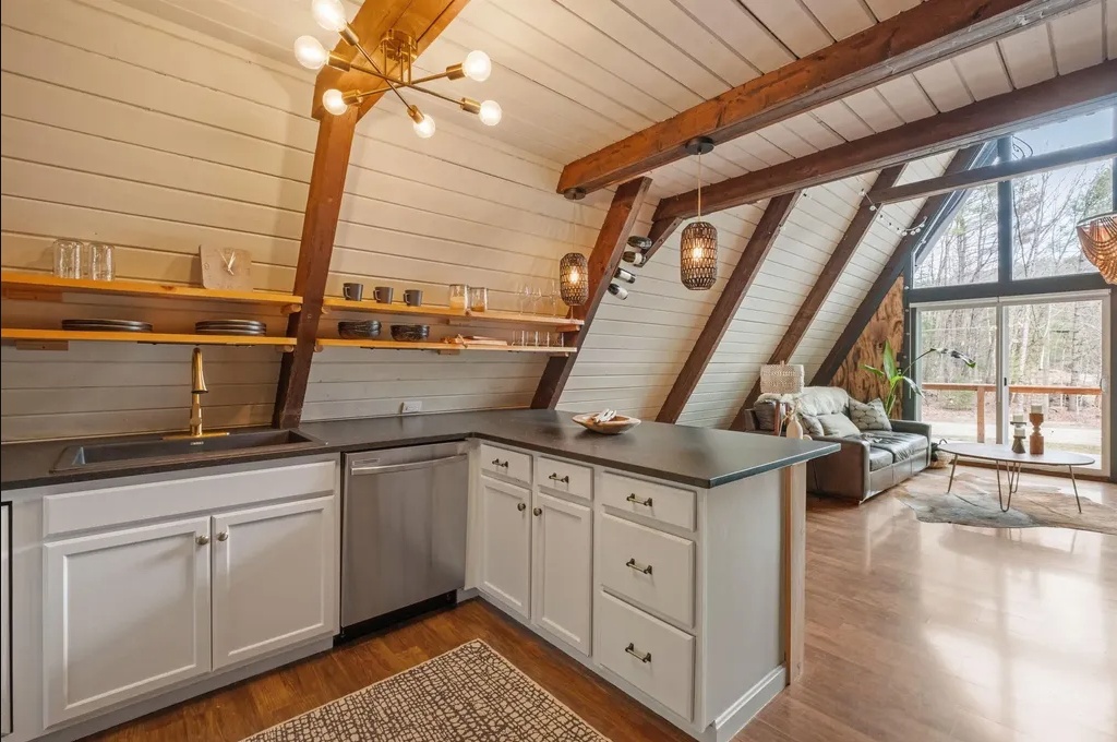 A modern A-frame kitchen with white cabinets, open wooden shelves, a dark countertop, and exposed wooden beams, adjacent to a living area with large windows.