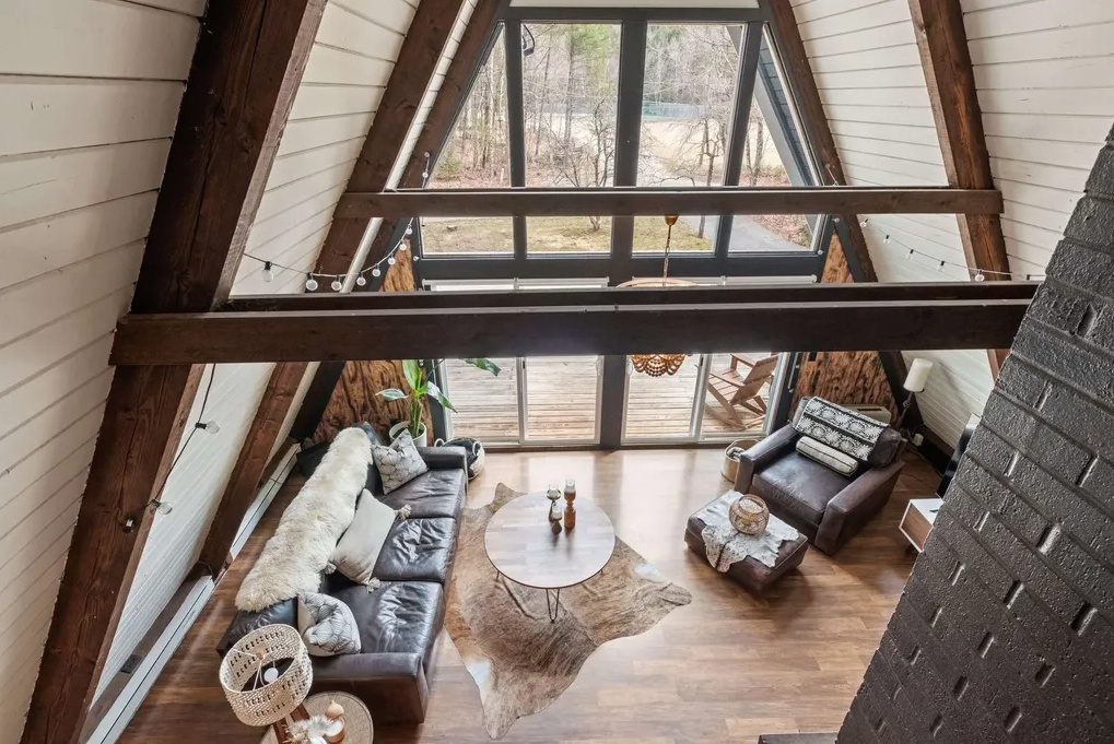 A-frame cabin living room with large windows, wooden floors, a fur-covered sofa, two armchairs, a round coffee table, and a cowhide rug, overlooking a wooded outdoor area.