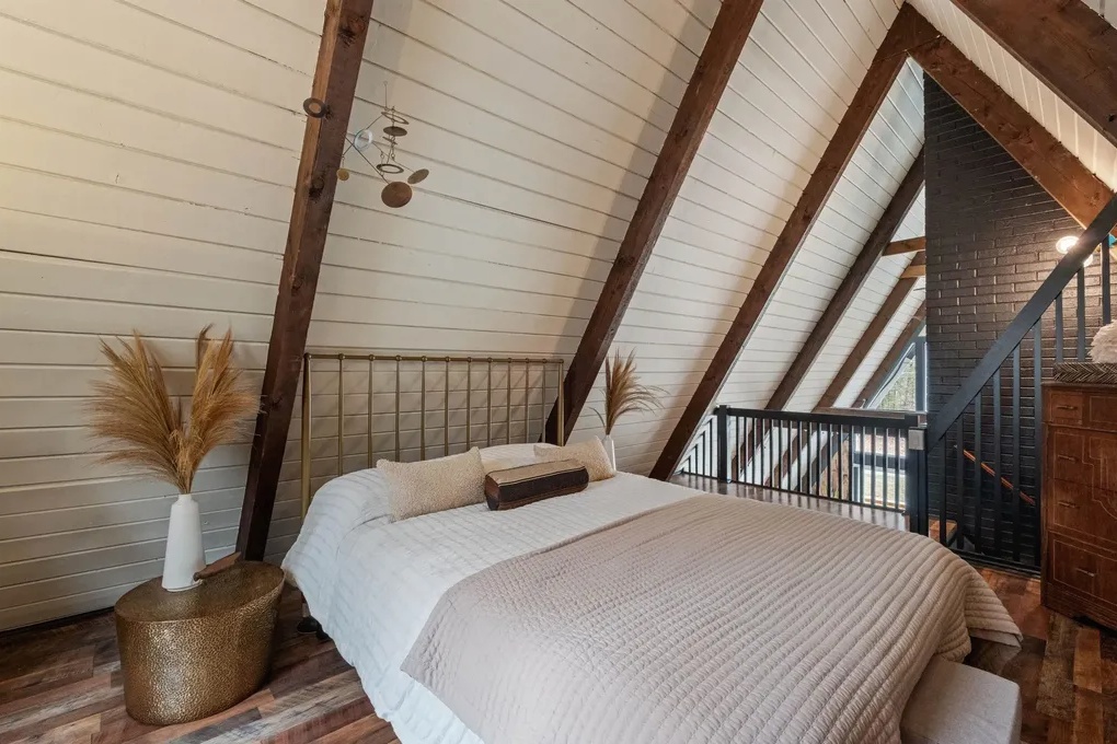 A cozy bedroom in an A-frame cabin features a large bed with neutral bedding, wooden beams, a gold side table, decorative pampas grass, and a black accent wall by the stairs.