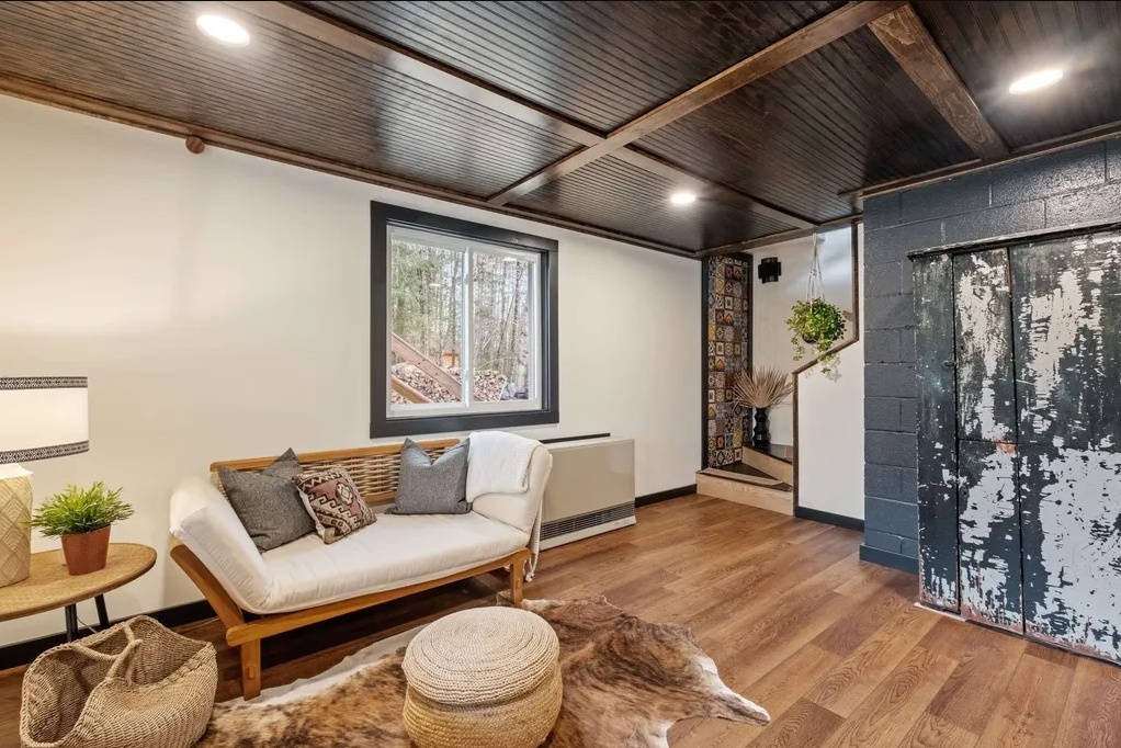 A cozy room with a wooden floor, white walls, a dark wood ceiling, a small sofa, a side table with a plant, a pouf, a cowhide rug, and a distressed sliding barn door.