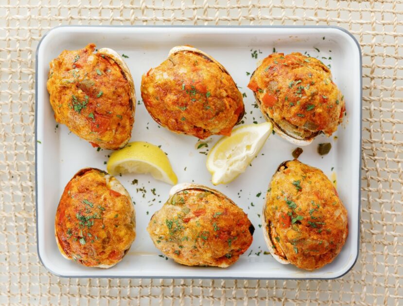 Six stuffed quahogs topped with herbs on a white tray, garnished with two lemon wedges, set on a woven surface.
