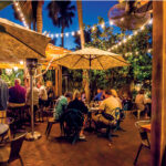 Third Wave restaurant, New Smyrna Beach. People sit at tables and under umbrellas in a warmly lit outdoor restaurant patio at dusk, with string lights overhead and trees in the background.