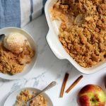 Apple crumble in a baking dish with a serving on a plate, topped with ice cream. Two whole apples, cinnamon sticks, and a striped cloth are nearby on a marble surface.