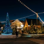A Christmas tree adorned with blue lights stands amidst buildings decorated with white lights. People walk on a path under string lights in a snowy setting at night.