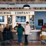 Customers standing at a brewery bar counter, with a staff member serving drinks. Menu boards are displayed on the wall behind them.