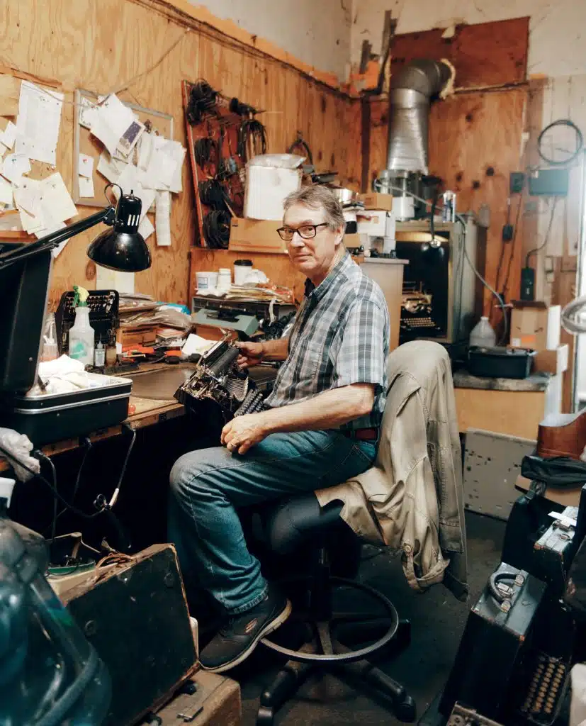 A person sits at a cluttered workbench in a workshop, surrounded by various tools, papers, and equipment.