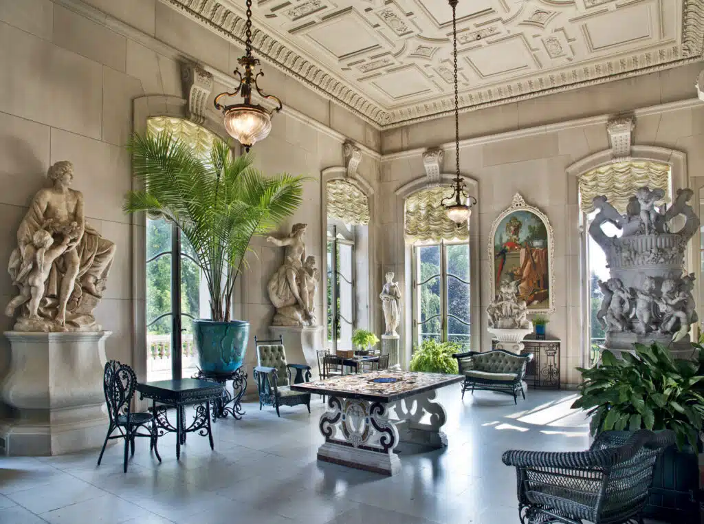Elegant sunroom with ornate ceiling, large windows, classical statues, potted plants, wrought iron furniture, and a mosaic-topped table. Natural light fills the space.